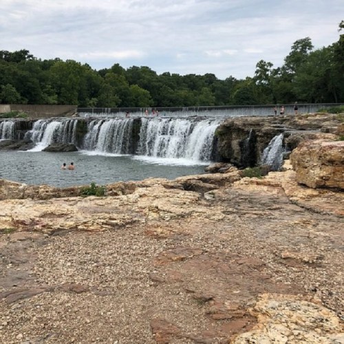 A scenic view of a waterfall with people swimming nearby, surrounded by lush greenery and rocky terrain.
