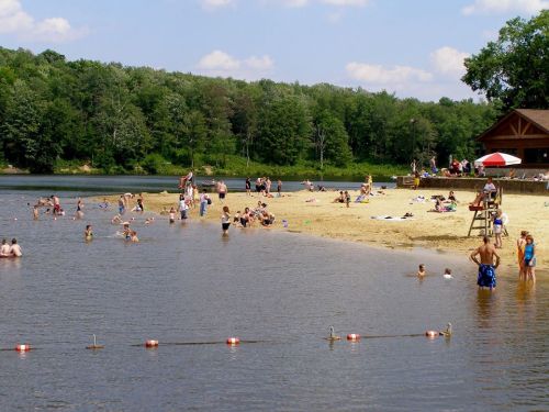 A sandy beach by a lake with people swimming, sunbathing, and enjoying the outdoors on a sunny day.