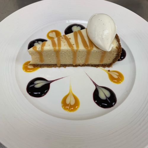 A slice of cheesecake on a plate, drizzled with caramel and berry sauces, topped with a dollop of whipped cream.