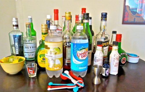 A variety of liquor bottles, mixers, and bar tools arranged on a table, with lime slices and a shot glass.