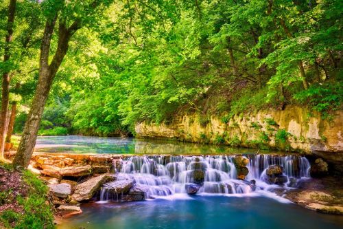 A serene waterfall cascades into a clear blue pool, surrounded by lush green trees and rocky cliffs.