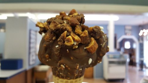 A close-up of a chocolate-dipped ice cream cone topped with crunchy bits, set in a bright indoor space.