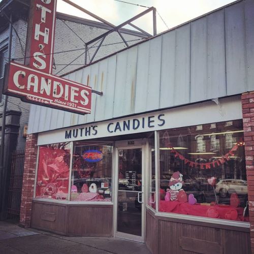 A storefront of Muth's Candies, featuring a vintage sign and a window display with pink decorations and candy.