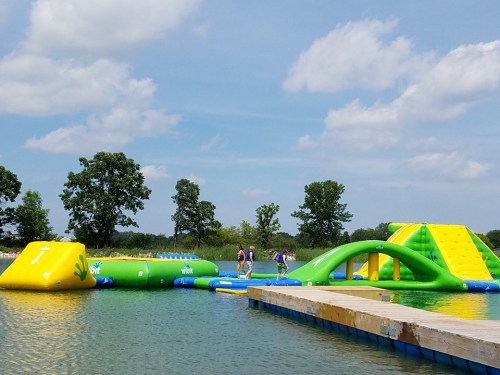 A colorful inflatable water park with slides and obstacles on a lake, surrounded by trees and a blue sky.