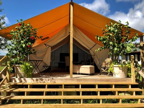 A glamping tent with an orange canopy, wooden steps, and potted plants, set against a blue sky with clouds.