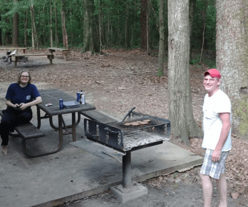 A man and a woman sit near a grill in a wooded picnic area, with picnic tables in the background.