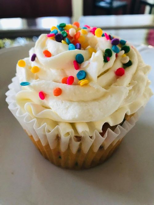 A colorful cupcake topped with swirls of white frosting and vibrant sprinkles on a white plate.
