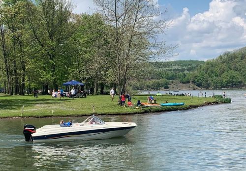 A boat glides on a lake near a grassy area with people picnicking and kayaking under a blue sky with fluffy clouds.