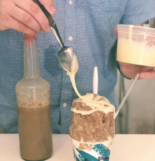A person drizzles a creamy sauce over a large ice cream treat, with a bottle of liquid nearby.