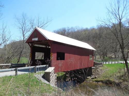 A red covered bridge spans a small stream, surrounded by green grass and bare trees under a clear blue sky.