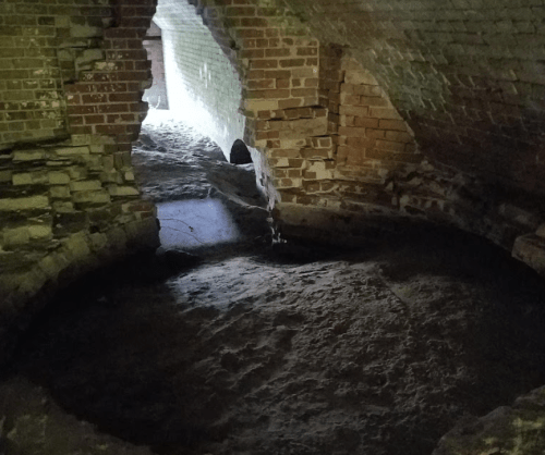 An underground space with brick walls and a dirt floor, featuring an arched opening and natural light coming through.