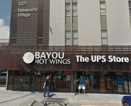 A storefront featuring Bayou Hot Wings and The UPS Store, with two people outside the entrance.