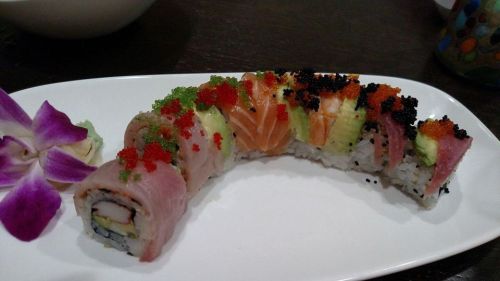 A colorful sushi roll topped with various fish, avocado, and fish roe, garnished with an orchid on a white plate.