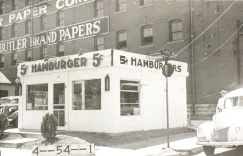 A vintage hamburger stand with a sign advertising 5-cent hamburgers, set against a backdrop of old buildings.