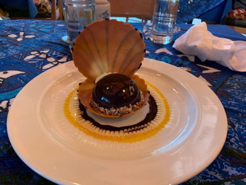 A dessert featuring a chocolate dome on a cookie base, surrounded by colorful sauces and a decorative shell.