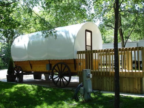 A vintage-style covered wagon on wheels, set on a grassy area with trees and a wooden deck nearby.