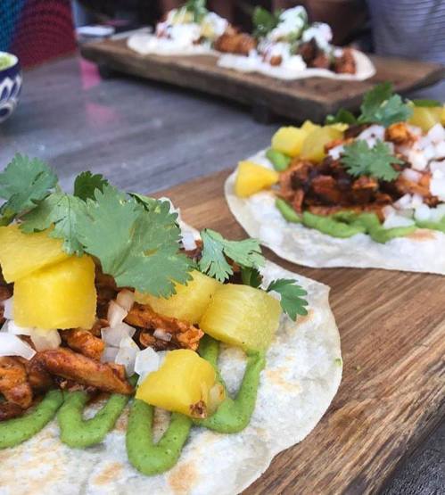 Two tacos on a wooden board, topped with pineapple, onions, cilantro, and a green sauce. Background features more tacos.