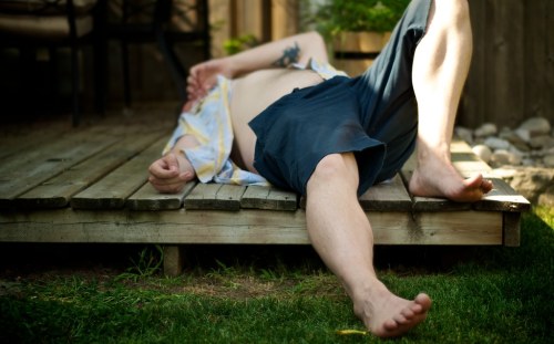 A person lies on a wooden deck, wearing shorts and a loose shirt, with one arm resting on their stomach.