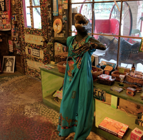 A quirky shop interior featuring a mannequin in a blue dress, resembling a dinosaur, surrounded by colorful decor and items.