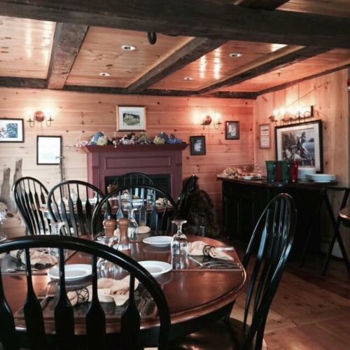 Cozy dining room with wooden walls, a fireplace, and a round table set for a meal. Warm, inviting atmosphere.