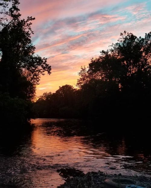 A serene river at sunset, with vibrant orange and pink skies reflecting on the water, surrounded by trees.