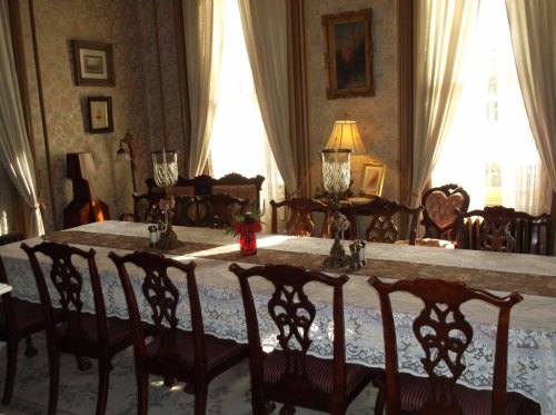 Elegant dining room with a long table, ornate chairs, and soft lighting from large windows. Decorated with vintage accents.