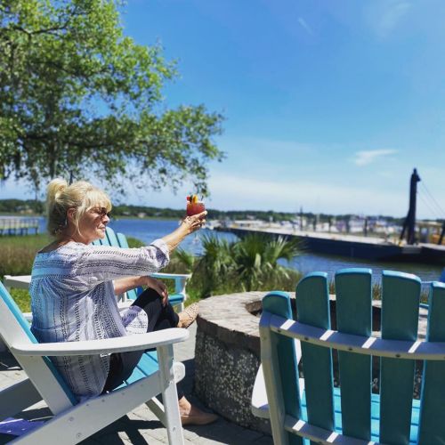 A woman enjoys a colorful drink while sitting in a chair by the water on a sunny day.