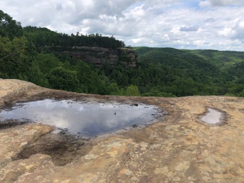 A rocky ledge with a small puddle reflects clouds above, overlooking a lush green valley and distant cliffs.