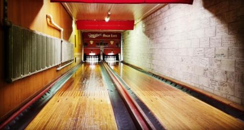 A view of a bowling alley lane with wooden flooring and illuminated pins at the end, surrounded by a brick wall.