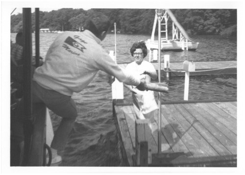 A man hands a fish to a woman on a dock, with a boat and diving platform in the background. Black and white photo.