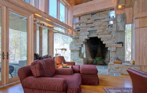 Cozy living room with large windows, a stone fireplace, and wooden accents, featuring plush seating and warm lighting.