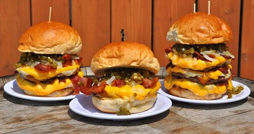 Three towering burgers with layers of cheese, bacon, pickles, and onions on toasted buns, served on white plates.
