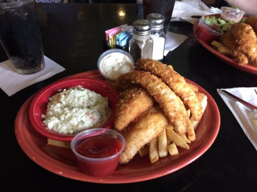 A plate of crispy fried fish fillets with fries, coleslaw, and dipping sauces on a black table.