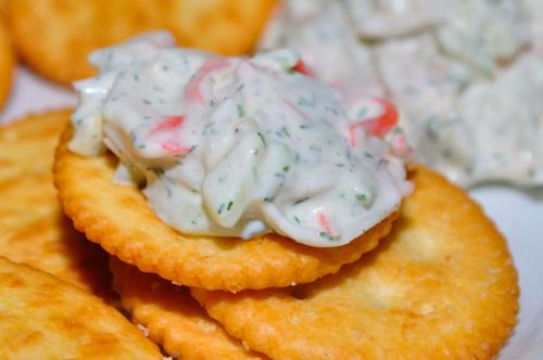 A close-up of a cracker topped with creamy dip, surrounded by more crackers and a bowl of dip in the background.