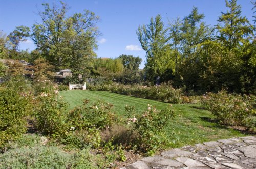 A lush garden with a manicured lawn, rose bushes, and trees under a clear blue sky. A stone path leads through the scene.