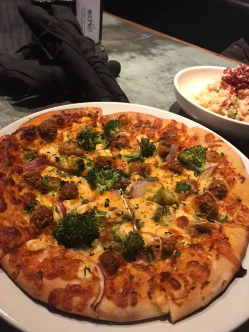 A pizza topped with broccoli, red onions, and meatballs, served on a plate with a side dish in the background.