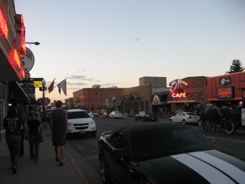 A street scene at dusk with pedestrians, storefronts, and a horse-drawn carriage in a small town.