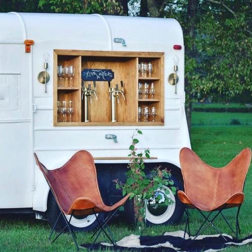 A vintage white trailer with a wooden bar, glasses, and two brown butterfly chairs in a grassy outdoor setting.