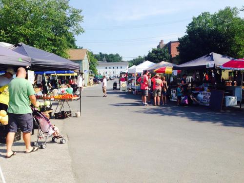 A bustling farmers market with vendors under tents, shoppers browsing fresh produce, and a sunny street scene.