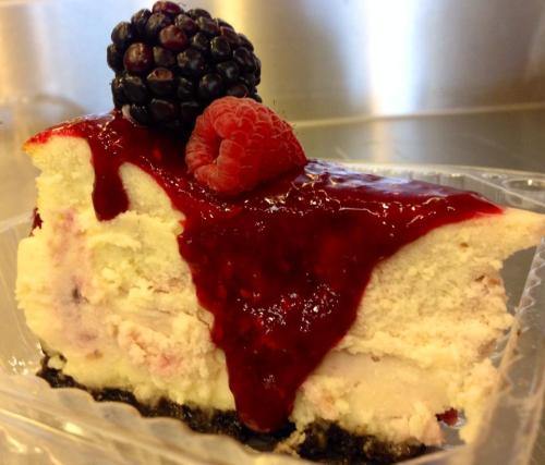 A slice of cheesecake topped with raspberry and blackberry, drizzled with red fruit sauce on a chocolate crust.