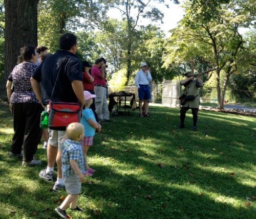 A group of people watches a demonstration in a park, with one person holding a rifle and others observing attentively.