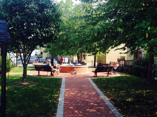A park scene with people sitting on benches around a circular area, surrounded by trees and greenery.