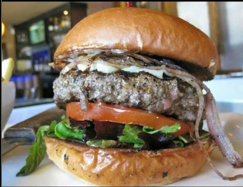 A close-up of a juicy burger with lettuce, tomato, and grilled onions on a toasted bun, served in a restaurant.