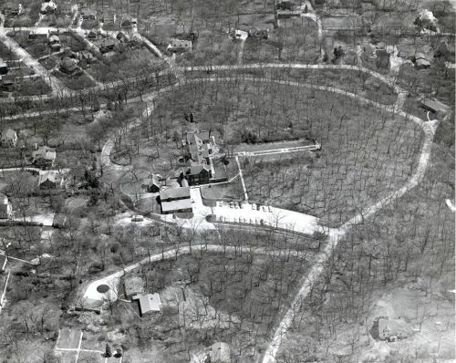 Aerial view of a large estate surrounded by trees and winding roads, with nearby residential areas visible.