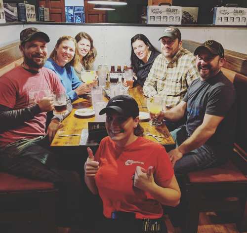 A group of seven people sitting at a table in a restaurant, with a waitress giving a thumbs up in front.