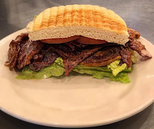 A large sandwich with layers of grilled meat, lettuce, and a toasted top bun on a white plate.