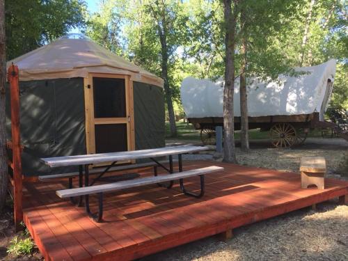 A yurt and a covered wagon are set in a wooded area, with a wooden deck and picnic table in front.