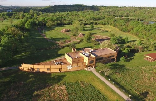 Aerial view of a modern building surrounded by lush greenery and open fields, with pathways and natural landscapes.