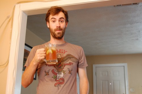 A man holding a mug of iced tea stands in a doorway, wearing a graphic t-shirt and looking playfully surprised.