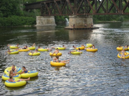 People float on colorful inner tubes in a river beneath a railway bridge, surrounded by lush greenery.